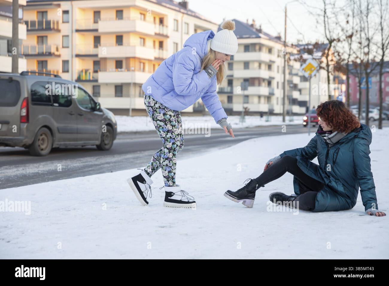 Due donne si trovano su un marciapiede non innevato vicino a una strada trafficata su cui viaggiano le auto. Una delle donne è seduta nella neve perché è caduta Foto Stock