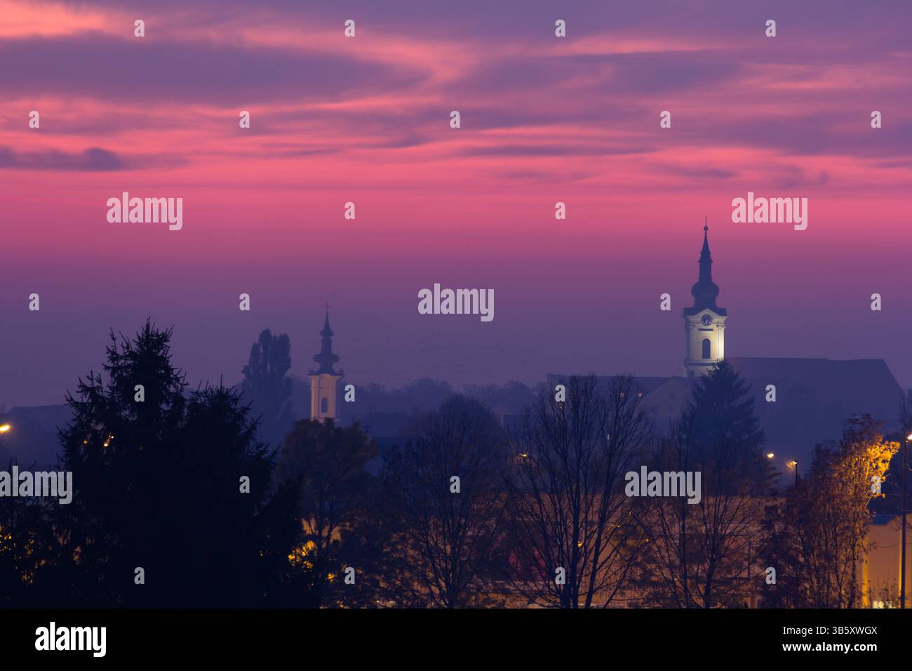 Tranquilla vista serale di un paesaggio urbano caratterizzato da torri e case della chiesa adagiate su un vibrante cielo al tramonto, evocando sentimenti di serenità e pace Foto Stock