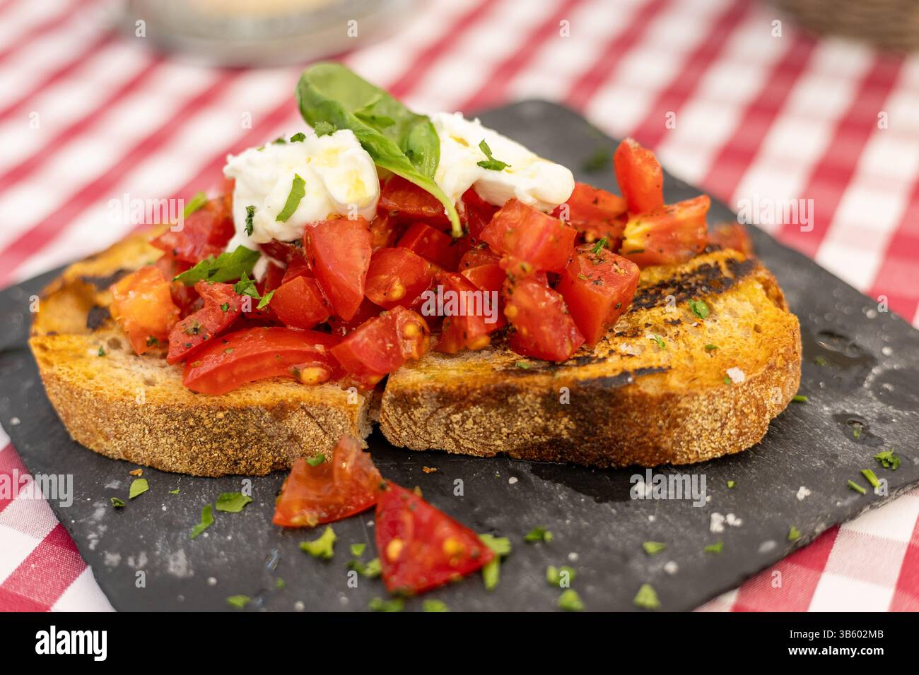 Il pane appena grigliato è ricoperto da pomodori rossi a dadini, formaggio cremoso e basilico profumato su un piatto nero testurizzato. Il pasto è impostato su una scheda contrassegnata Foto Stock