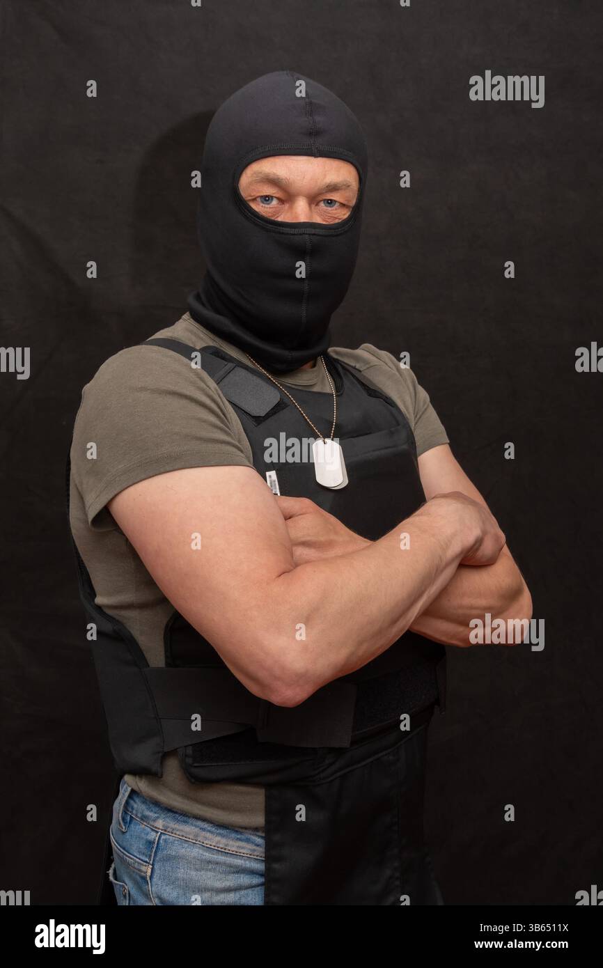Un uomo che indossa una balaclava e un'armatura per il corpo poggia su uno sfondo scuro con le braccia incrociate Foto Stock