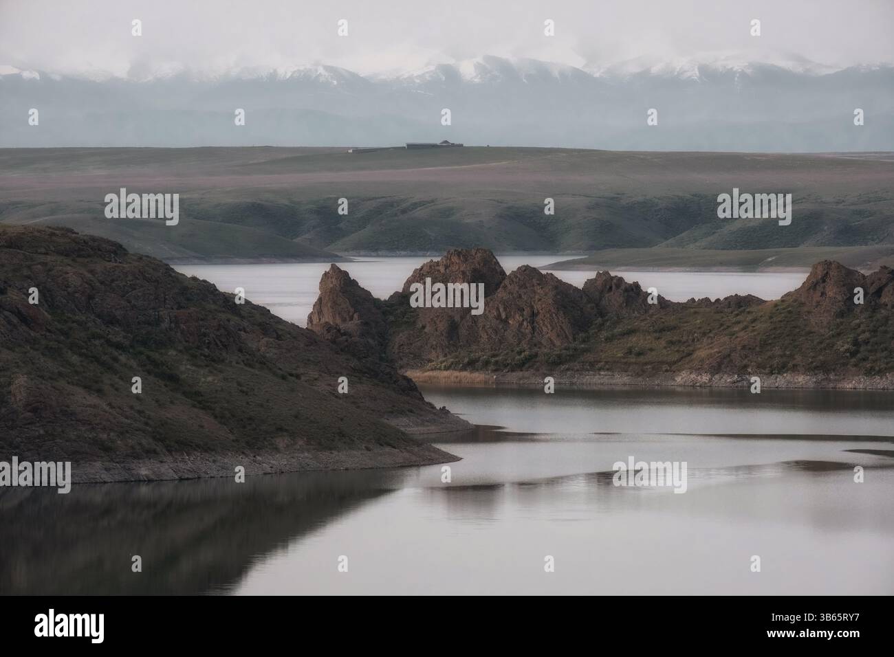 I suggestivi affioramenti rocciosi sorgono dalla riva del lago artificiale Kurtinskoye in Kazakistan, con lontane montagne innevate visibili all'orizzonte. Un tranq Foto Stock