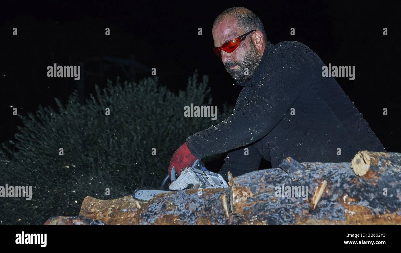 Un uomo con occhiali protettivi e barba lavora di notte con una motosega su una pila di legno, Psinthos del fuoco pasquale, sabato Santo, Pasqua, Psinthos, Rhode Foto Stock