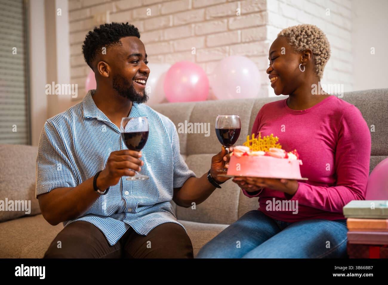 Felice giovane coppia che festeggia il compleanno della donna a casa. La donna tiene la torta e brindano con il vino. Foto Stock