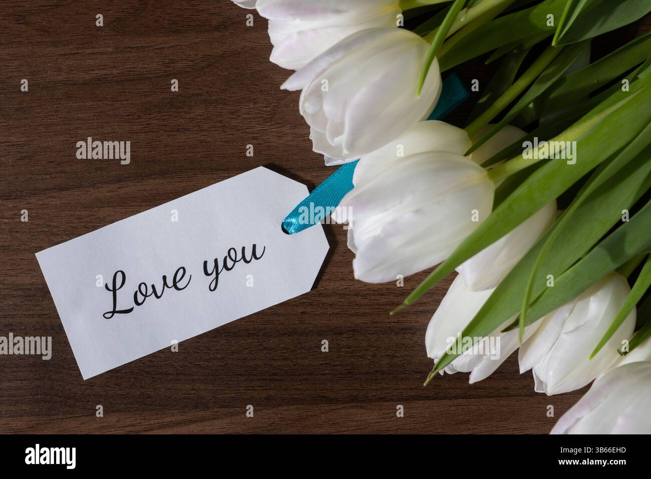 Eleganti tulipani bianchi disposti accanto a un tag che mostra il testo Love You su una superficie in legno scuro, perfetti per esprimere affetto d'amore e messaggio romantico Foto Stock