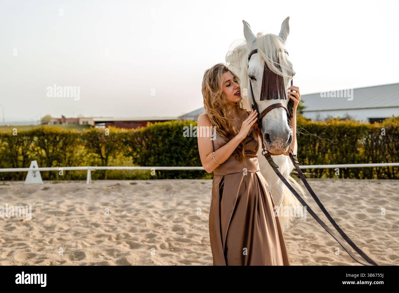 Elegante giovane donna in un abito fluente che interagisce con tenerezza con un cavallo bianco in un paddock di sabbia. Foto Stock