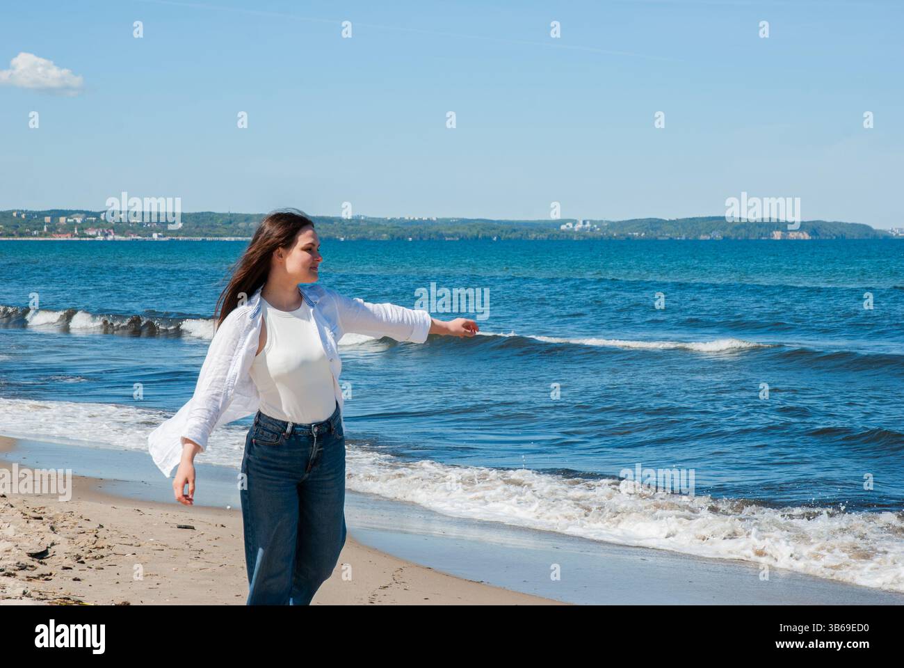 Giovane donna caucasica che si gode la spiaggia soleggiata con le onde e il cielo azzurro che cammina lungo la riva in jeans casual e abbigliamento bianco. Digitale Foto Stock