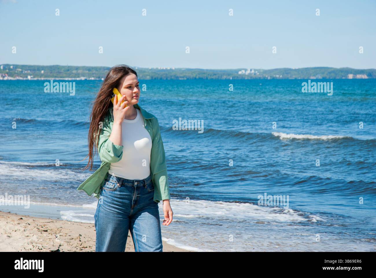 Giovane donna caucasica che cammina lungo l'oceano in una giornata di sole, parla su uno smartphone, mostra moda casual da spiaggia e momenti spensierati. Digitale nom Foto Stock
