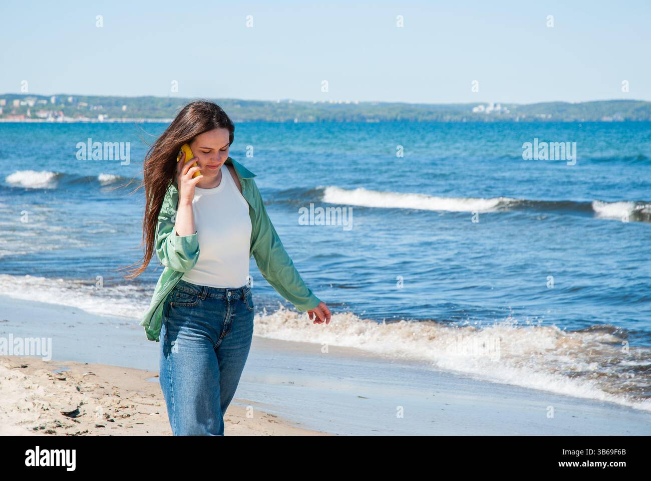 Giovane donna caucasica che cammina lungo la spiaggia mentre parla al telefono, indossa una camicia verde e jeans, con le onde dell'oceano sullo sfondo. Digi Foto Stock