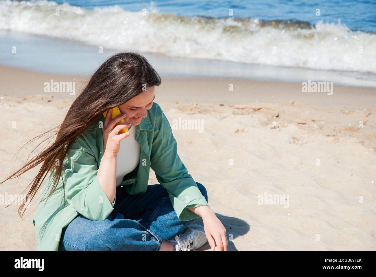 Giovane donna caucasica che si gode una giornata di sole in spiaggia mentre parla al telefono, seduto sulla sabbia vicino alle onde dell'oceano in una giacca verde informale. Digitale Foto Stock
