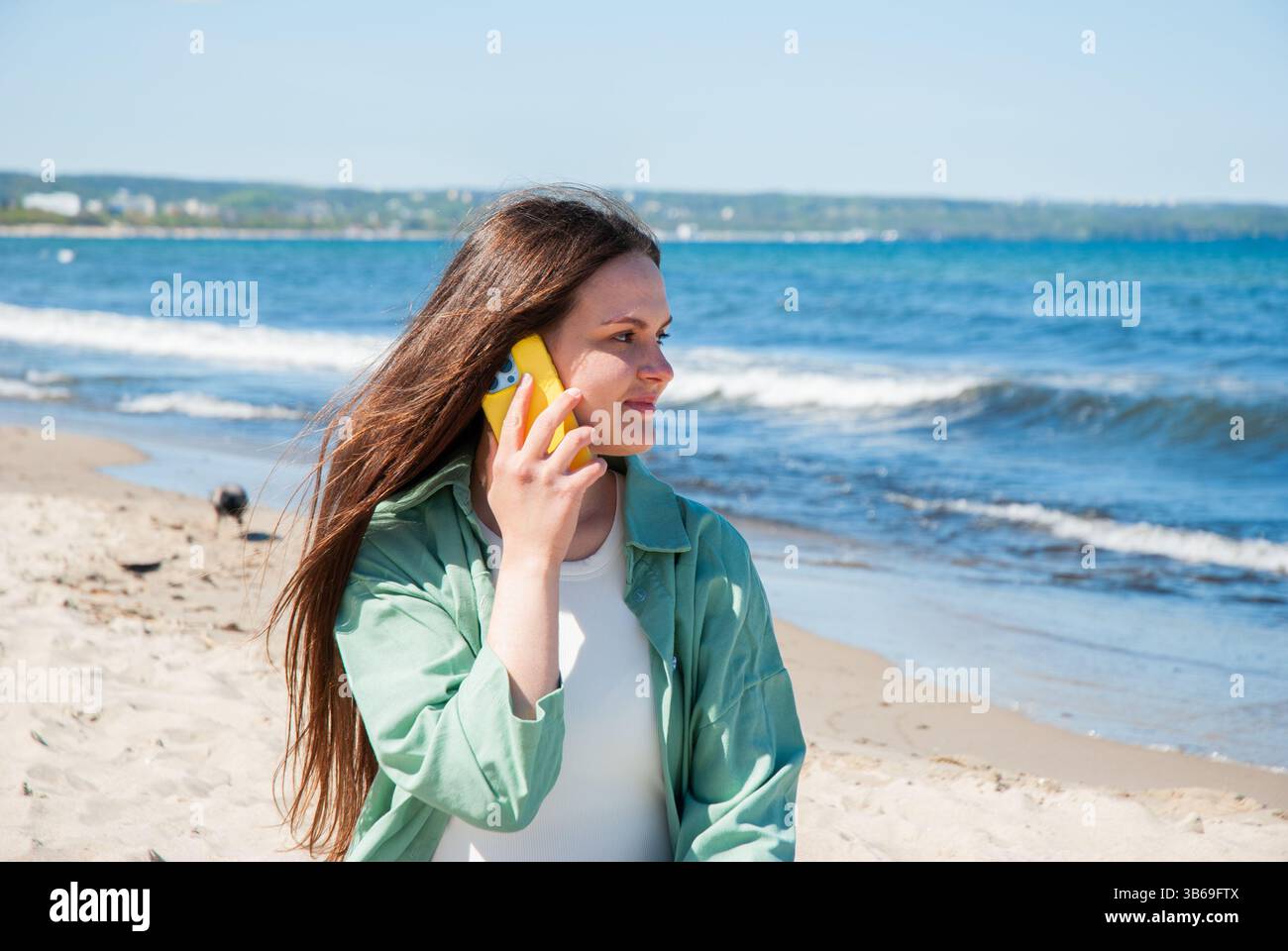 Giovane donna caucasica che si gode una giornata di sole sulla spiaggia mentre parla su uno smartphone con le onde e il cielo blu sullo sfondo. Stile di vita digitale nomade. Rif Foto Stock