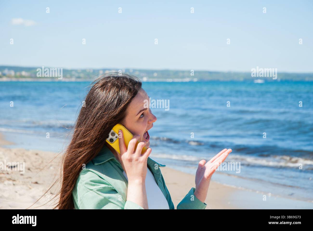 Giovane donna caucasica con lunghi capelli castani impegnata in una conversazione telefonica animata sulla spiaggia soleggiata, esprimendo una sorpresa con lo sfondo blu dell'oceano. Cifra Foto Stock