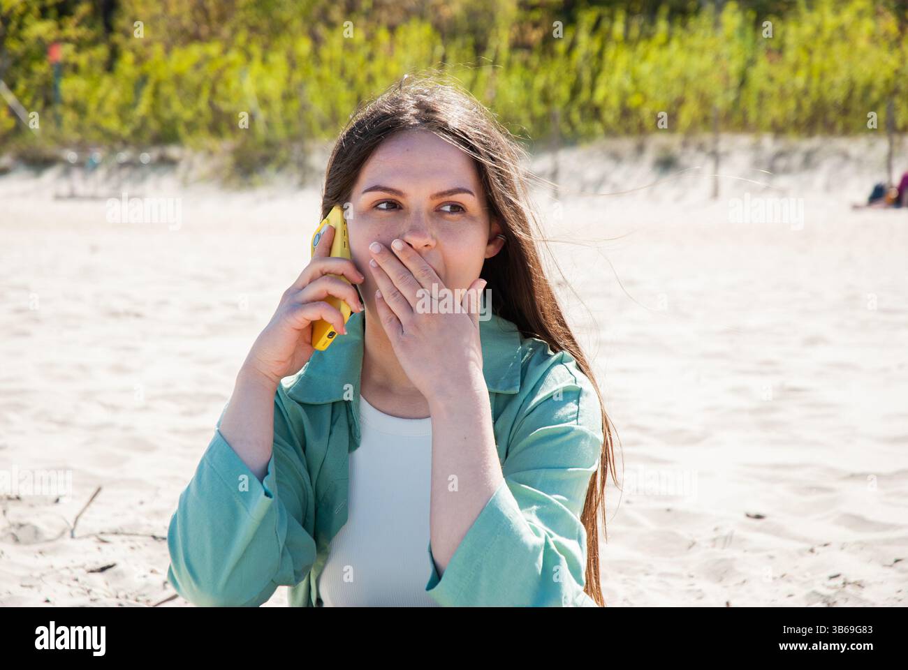 La giovane donna caucasica in spiaggia con una giacca verde si copre la bocca mentre parla con un telefono giallo, esprimendo sorpresa e contemplazione. Digitale Foto Stock