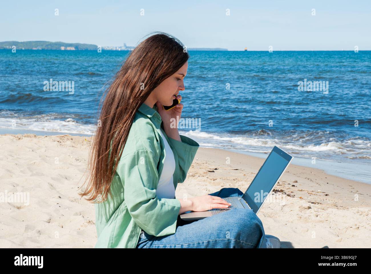 Giovane donna caucasica adulta con lunghi capelli castani siede sulla spiaggia di sabbia usando un computer portatile mentre parla al telefono vicino all'oceano blu sotto il cielo limpido. Digitale nom Foto Stock