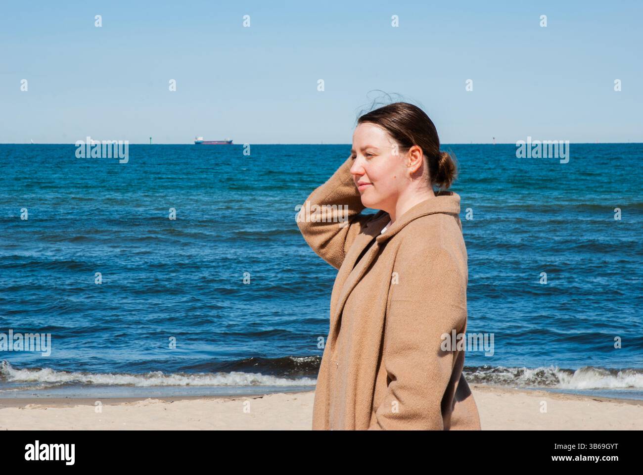 Donna adulta contemplativa caucasica che si gode una tranquilla giornata in spiaggia con un cielo azzurro e un oceano calmo sullo sfondo. Stile di vita dei nomadi digitali Foto Stock