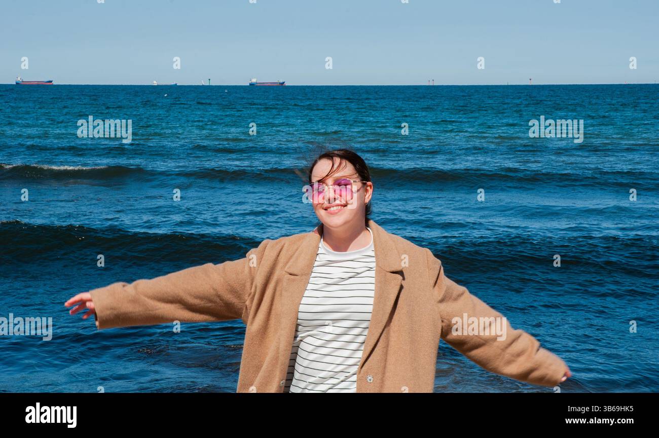 Giovane donna caucasica che si gode una giornata di sole sulla spiaggia, indossa uno stemma marrone, con le braccia allungate e occhiali da sole rosa, onde dell'oceano sullo sfondo Foto Stock