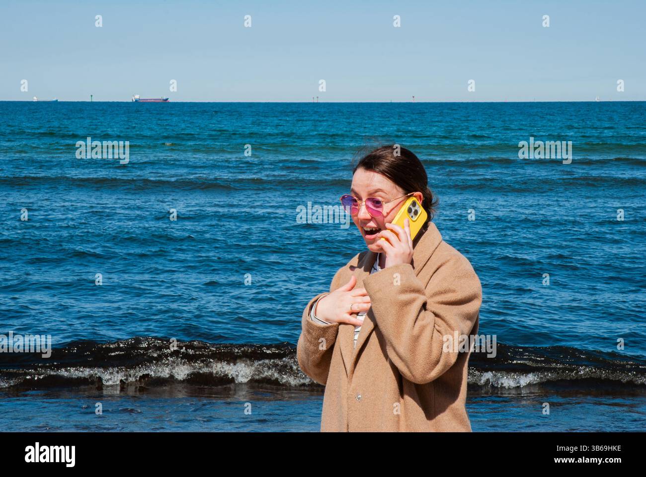 Giovane donna caucasica che parla su un telefono giallo in spiaggia, indossa occhiali da sole rosa e un cappotto marrone, con l'oceano sullo sfondo. Digitale nom Foto Stock