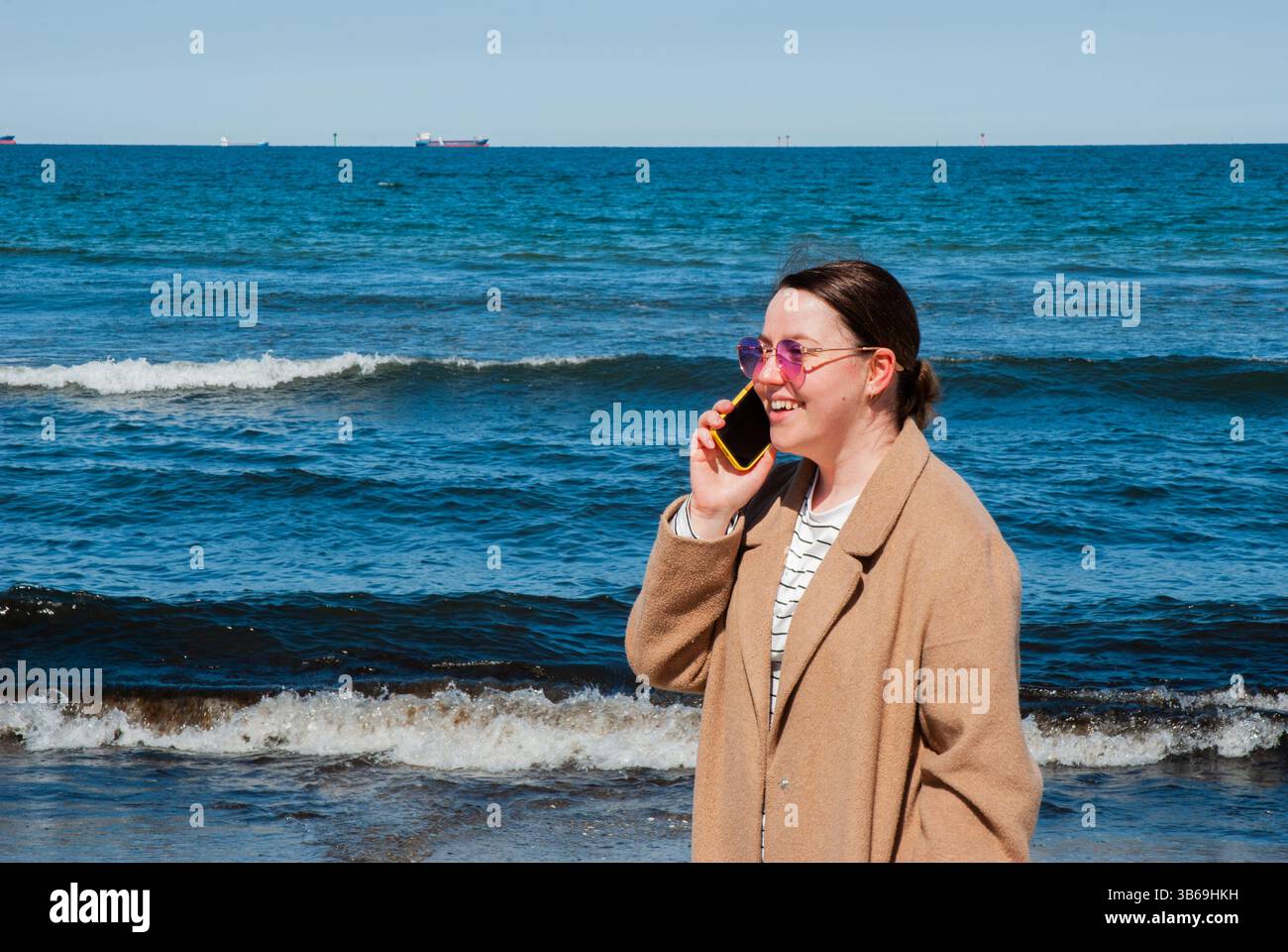 Giovane donna caucasica con un cappotto marrone che parla su uno smartphone in spiaggia con onde blu dell'oceano sullo sfondo in una giornata di sole. Nomadi digitali Foto Stock