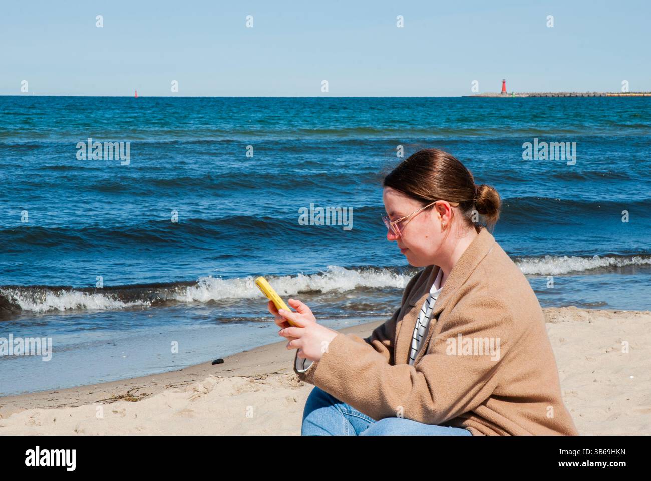 Giovane donna caucasica in un abbigliamento casual che si gode una giornata di sole sulla spiaggia mentre naviga sul suo smartphone, con le onde dell'oceano sullo sfondo. Digitale no Foto Stock