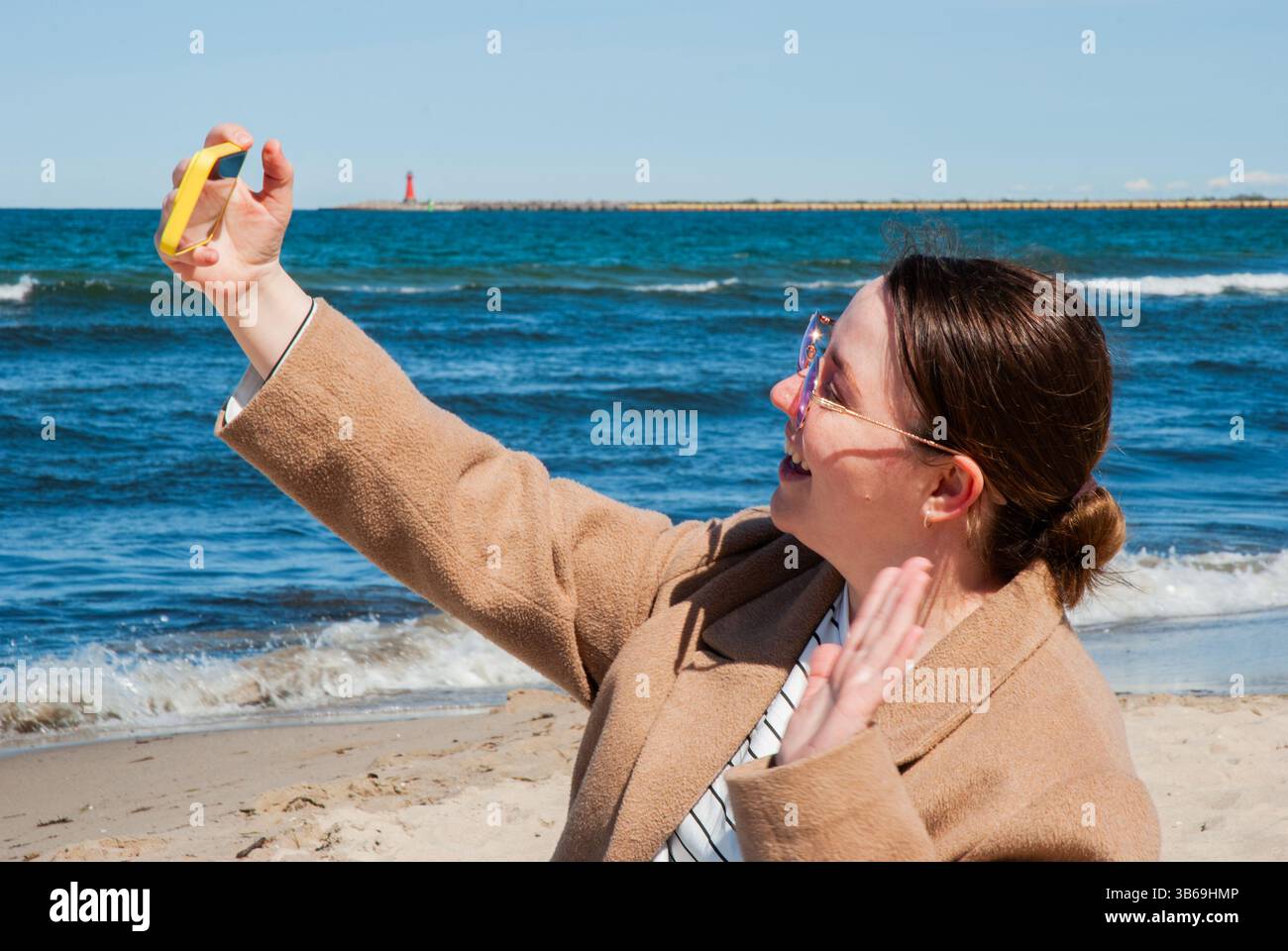Una giovane donna caucasica che si gode di una giornata di sole sulla spiaggia scatta selfie con la fotocamera gialla contro il vivace oceano blu e il faro in lontananza. Digitale nom Foto Stock