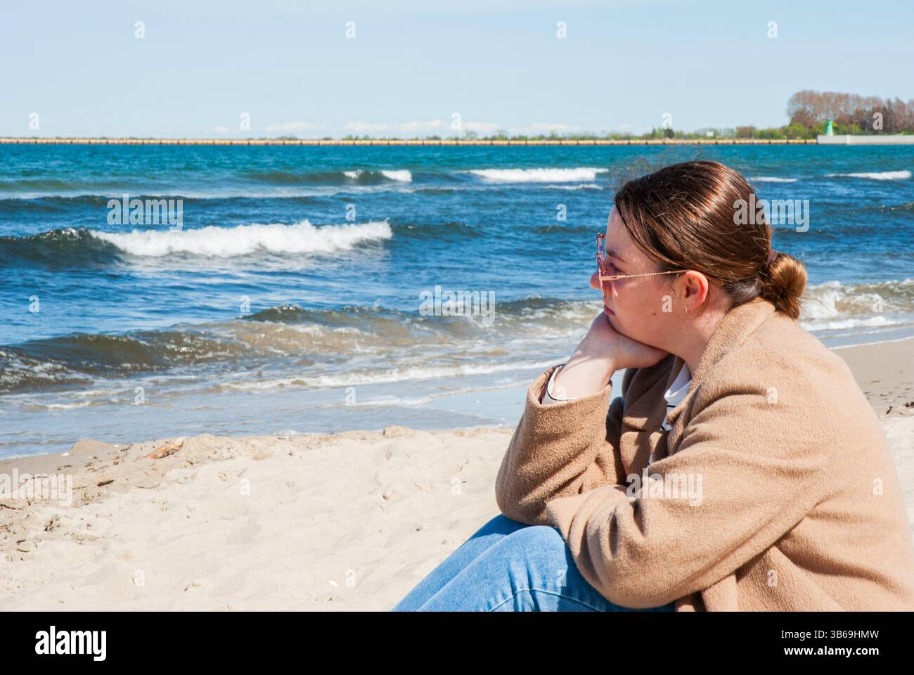 Giovane e premurosa donna caucasica seduta accanto all'oceano in una giornata di sole, contemplando, con le onde che si infrangono dolcemente su una spiaggia sabbiosa. Digitale no Foto Stock
