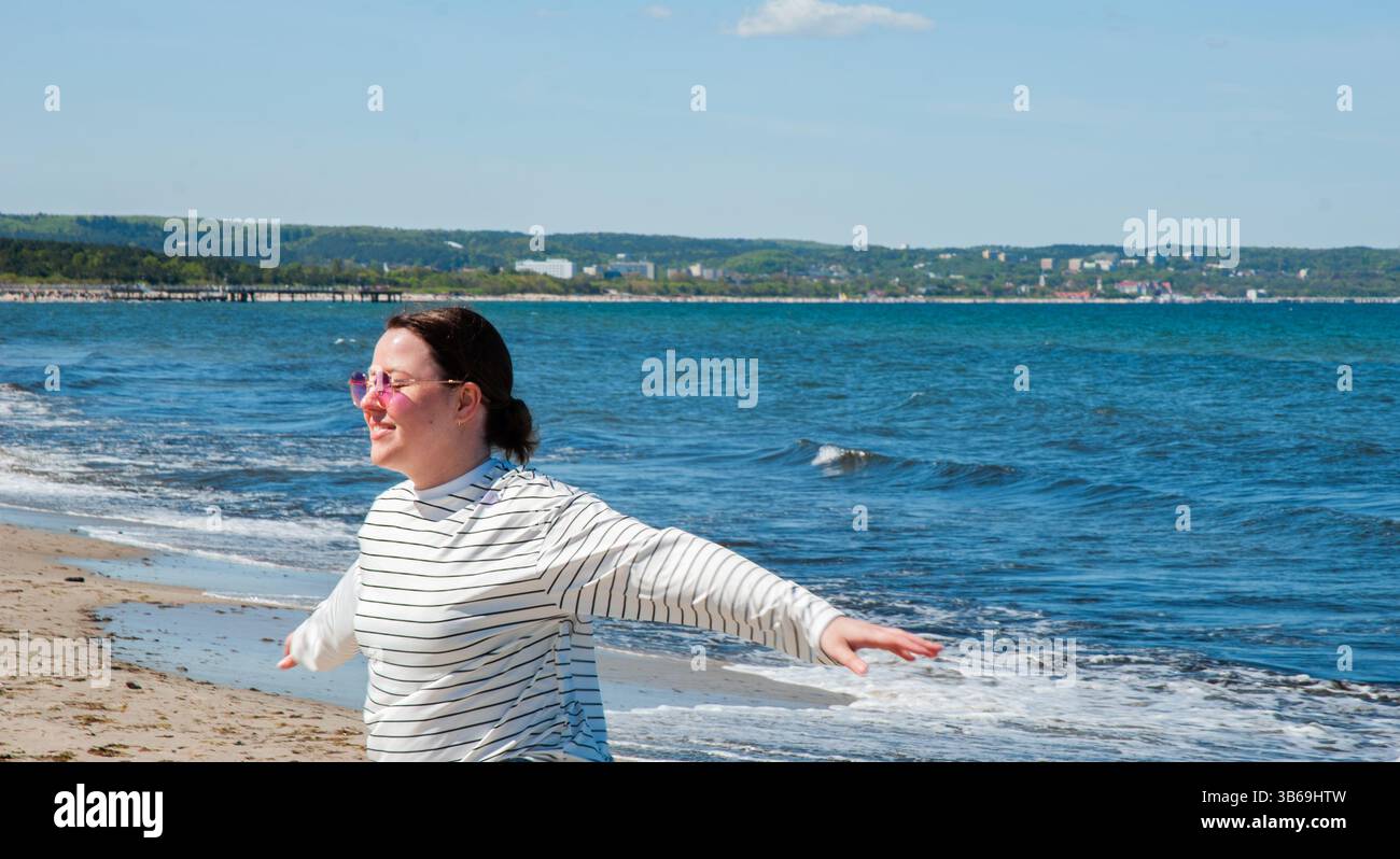 Giovane donna caucasica che si gode una giornata di sole sulla spiaggia, indossando occhiali da sole rosa e sorridendo ampiamente, circondata da onde e costa panoramica. Digita Foto Stock