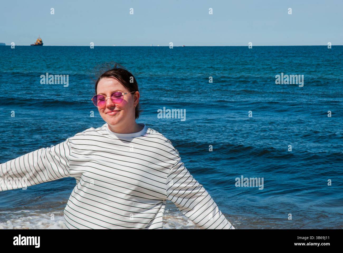 Giovane donna caucasica con maglione a righe e occhiali da sole che si gode una giornata di sole sulla spiaggia con le onde blu dell'oceano che creano uno sfondo sereno. Foto Stock