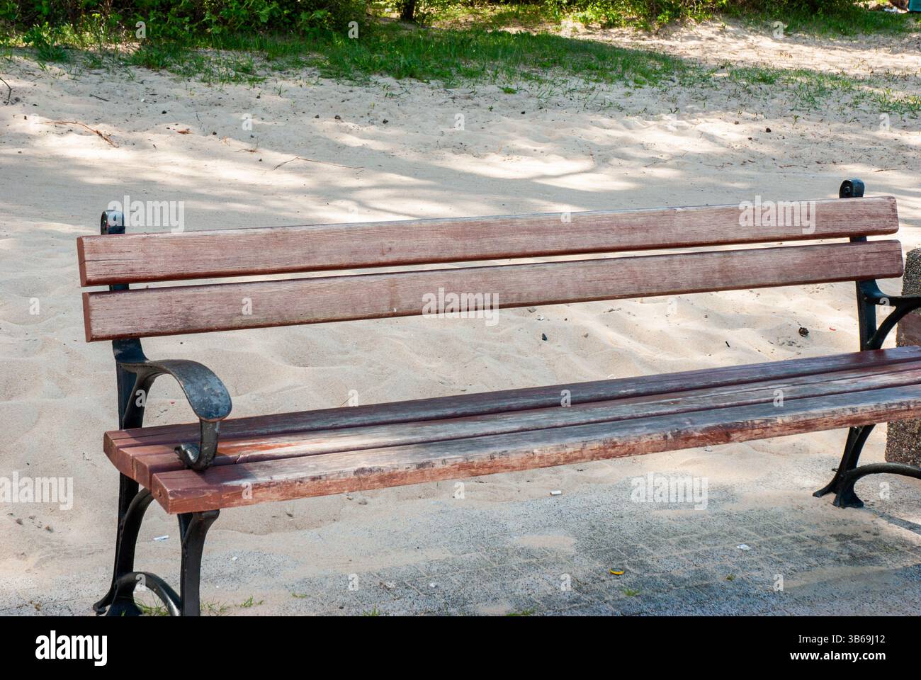Panchina in legno intemprata su un sentiero di sabbia con eleganti dettagli in ferro nel parco illuminato dal sole circondato da lussureggiante vegetazione e ombre che creano serenità. Digitale Foto Stock
