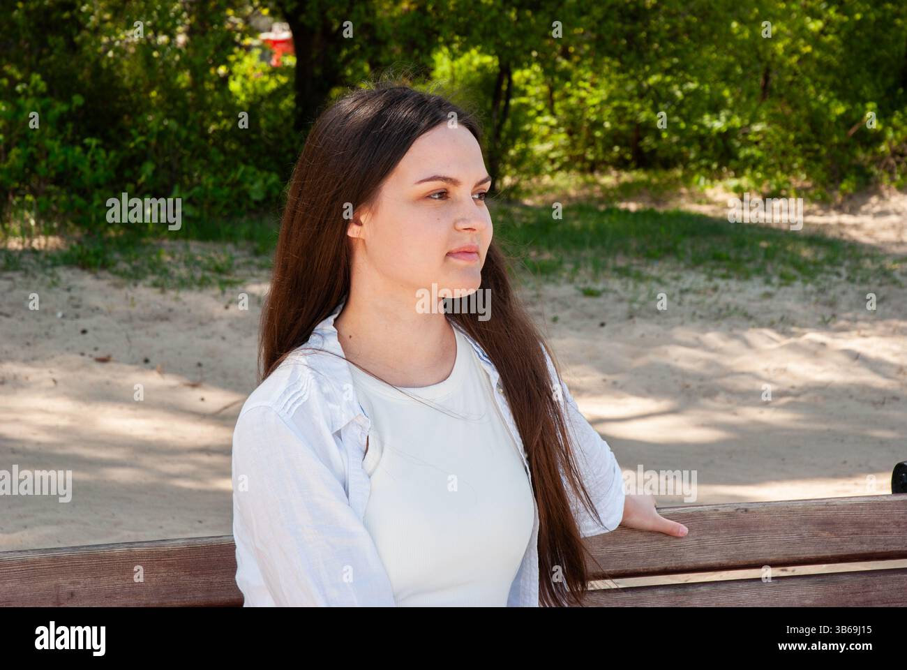 Giovane donna caucasica in camicia bianca con lunghi capelli marroni siede pensiamente su una panchina del parco circondata da verde e sabbia. Stile di vita digitale nomade. Foto Stock