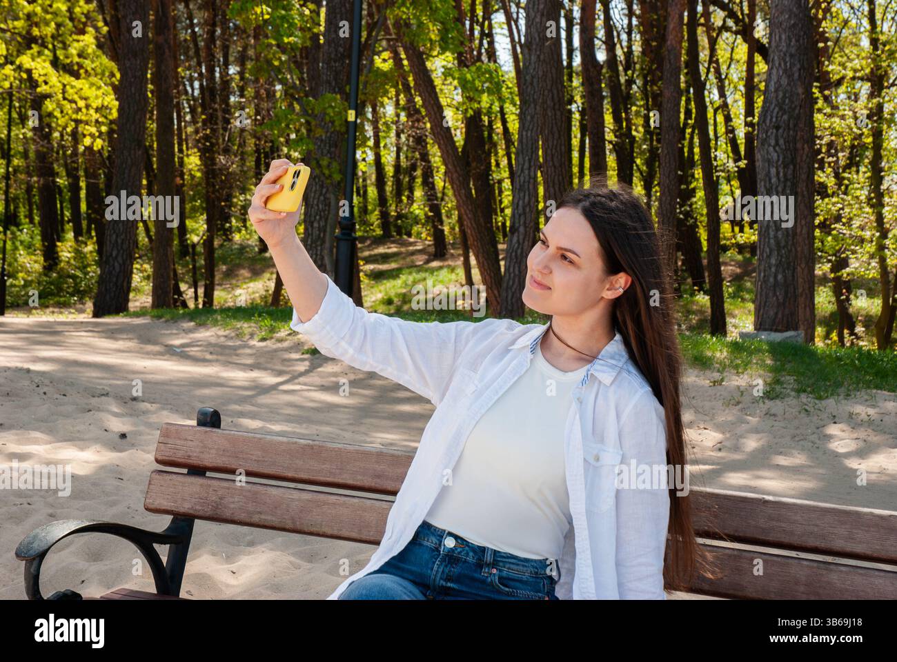 Giovane donna caucasica che fa un selfie in una giornata di sole in un parco, seduta su una panchina di legno circondata da alberi verdi e terreno sabbioso. Digitale Foto Stock