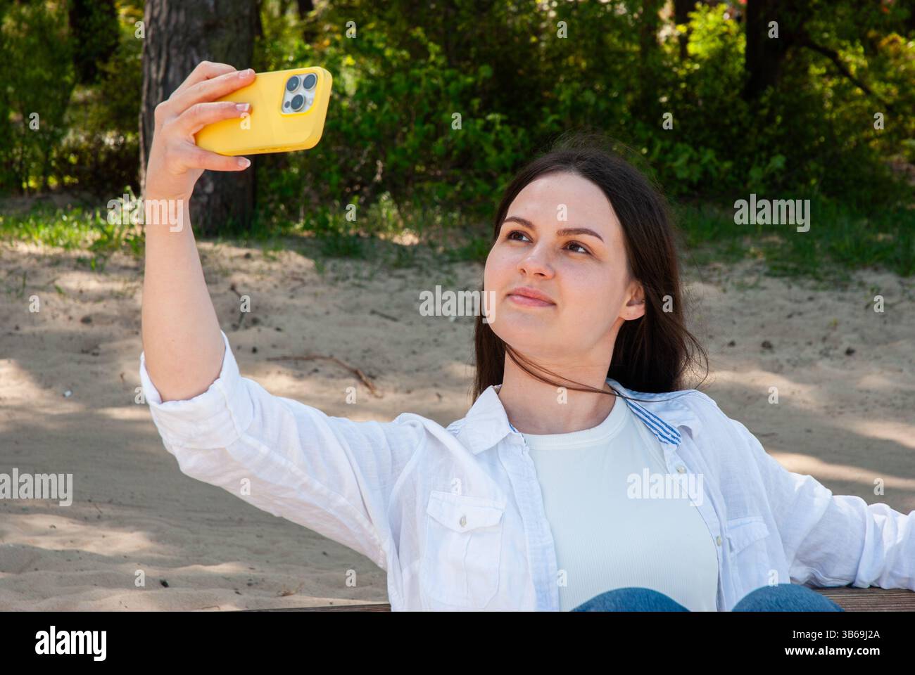 Giovane donna caucasica che scatta selfie in natura con lo smartphone giallo, indossa una giacca bianca informale e goditi una giornata di sole all'aperto. Nomad digitale li Foto Stock