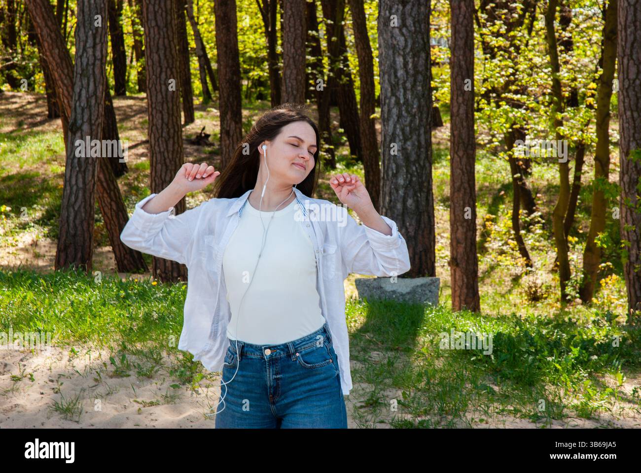 Giovane donna caucasica che ascolta la musica all'aperto in un ambiente tranquillo nella foresta, indossando un abbigliamento casual con un'espressione rilassata e auricolari. Nomade digitale Foto Stock
