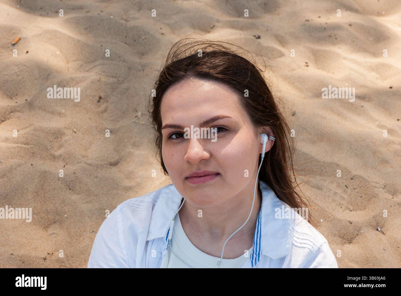 Giovane donna caucasica con auricolari sulla spiaggia, impegnata in momenti rilassanti, indossa un abbigliamento casual, circondata da spiagge sabbiose. Nomadi digitali Foto Stock