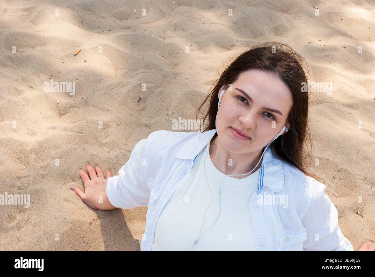 Giovane donna caucasica che si rilassa sulla spiaggia di sabbia ascoltando musica, godendosi una giornata di sole in camicia bianca informale con auricolari. Stile di vita digitale nomade. Foto Stock