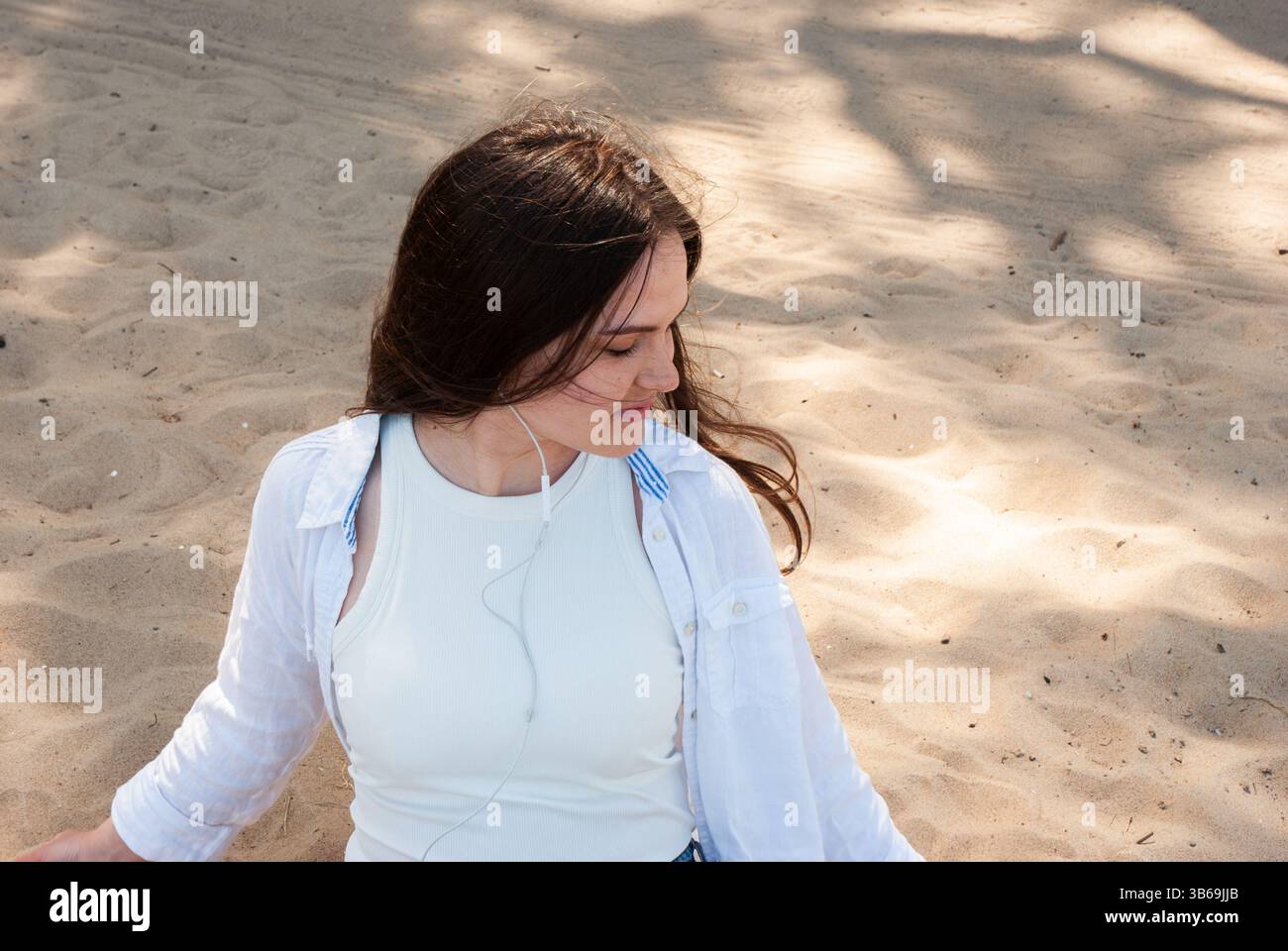 Giovane donna caucasica con lunghi capelli castani che si gode una giornata di sole sulla spiaggia di sabbia, indossa un abbigliamento casual bianco e ascolta musica. Nomade digitale Foto Stock