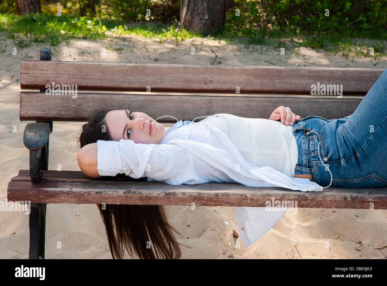Giovane donna caucasica in un abbigliamento informale che si rilassa su una panchina di legno in una spiaggia sabbiosa in una giornata di sole con la natura sullo sfondo. Il lifest dei nomadi digitali Foto Stock