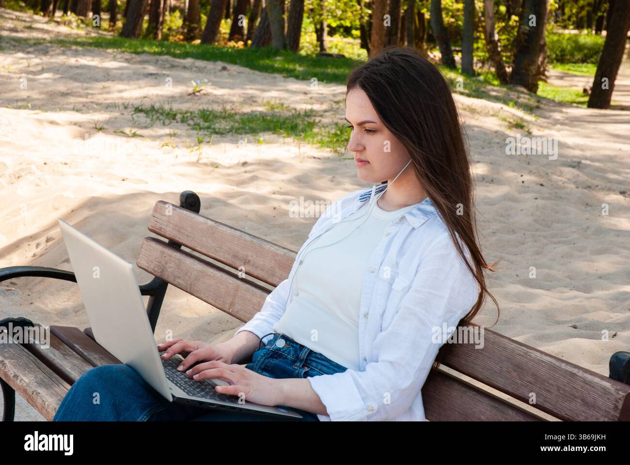 Giovane donna caucasica che usa un laptop su una panca soleggiata con auricolari, multitasking all'aperto su un tranquillo sentiero sabbioso tra lussureggianti alberi verdi. Digitale Foto Stock