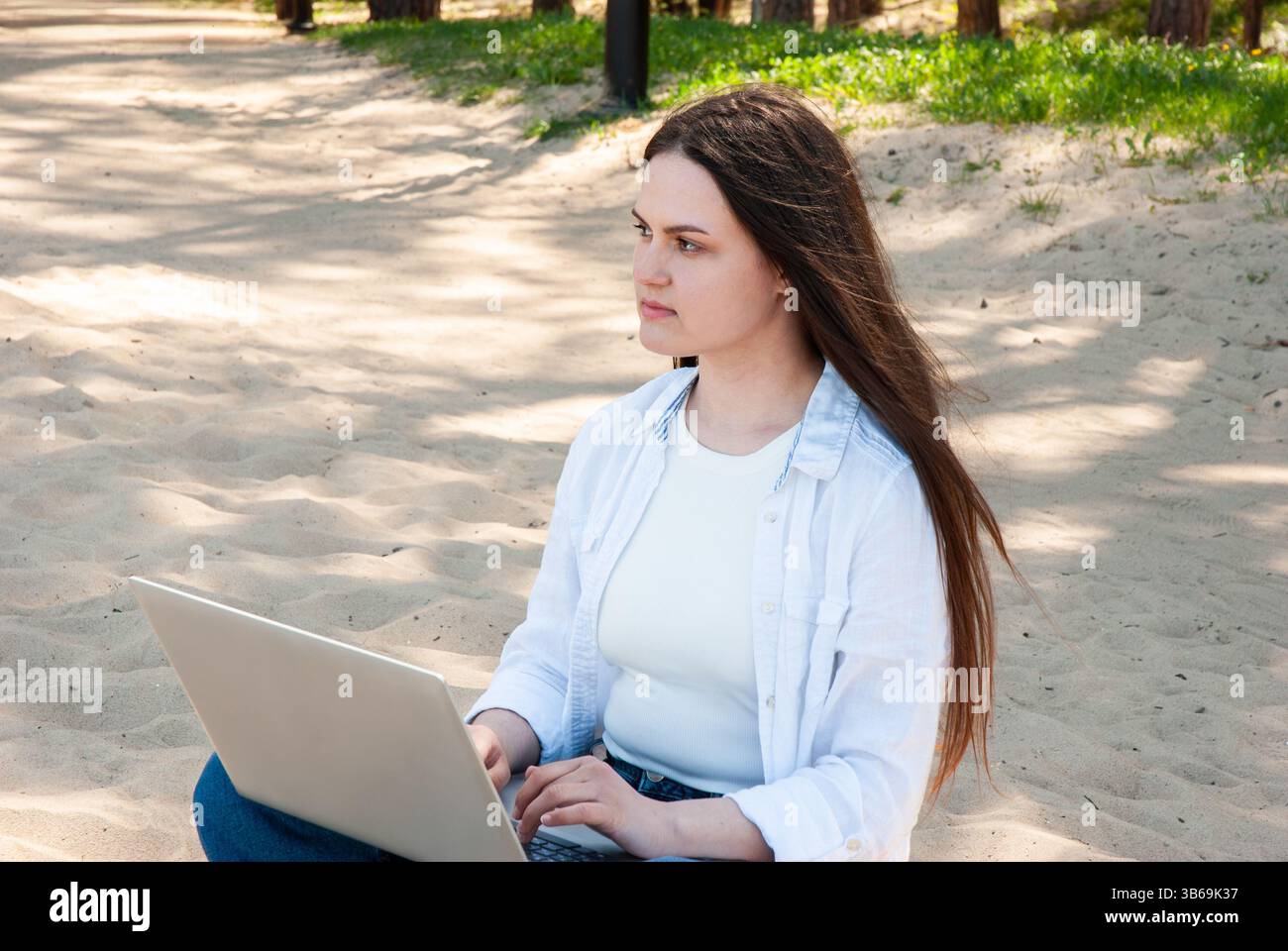 Giovane donna caucasica adulta con lunghi capelli castani su una spiaggia sabbiosa circondata da alberi e crogiolata alla luce naturale del sole. Nomad digitale Foto Stock