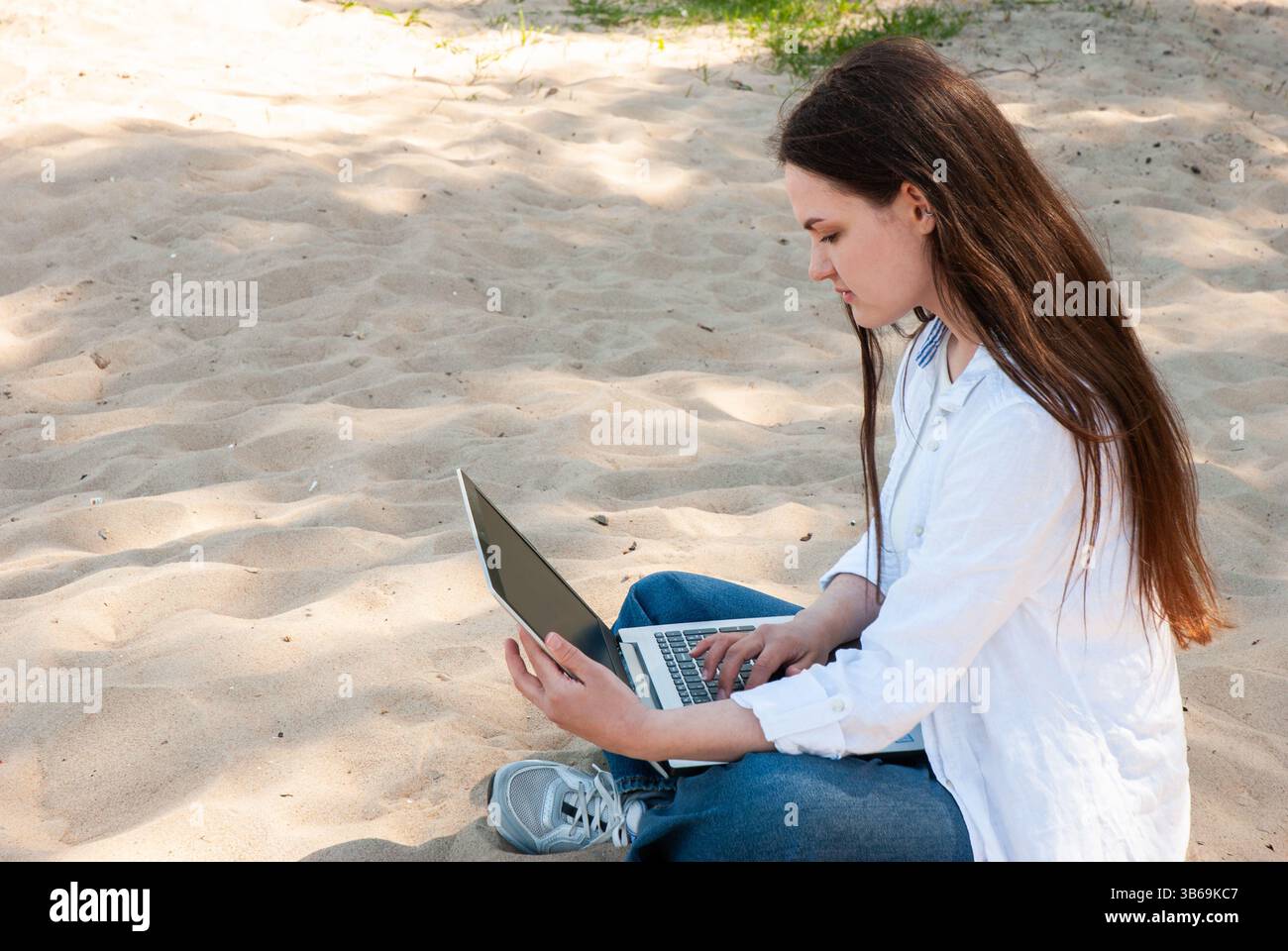 Giovane donna caucasica con lunghi capelli marroni che utilizza un computer portatile mentre si siede sulla spiaggia di sabbia, indossa camicia bianca e jeans casual, concentrandosi con attenzione. Digi Foto Stock