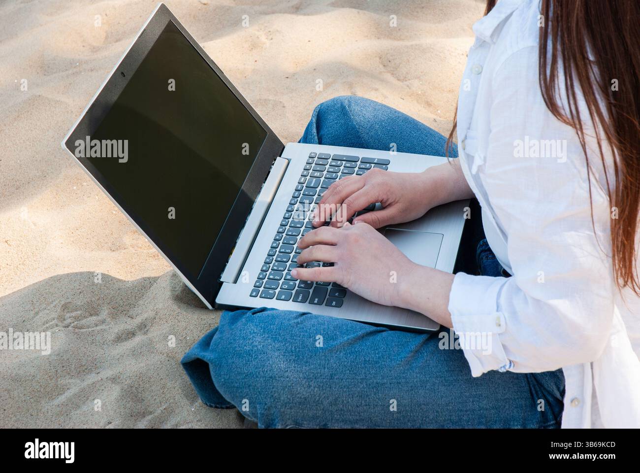 Giovane donna adulta che utilizza un computer portatile su una spiaggia sabbiosa e indossa una camicia bianca e un jeans, illustrando il lavoro a distanza e il relax in un ambiente costiero. Digi Foto Stock