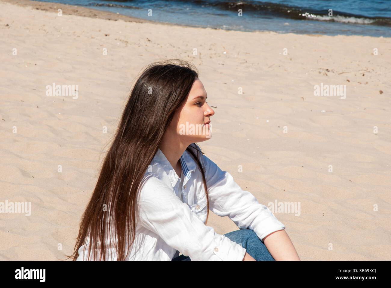 Giovane donna caucasica che si rilassa su una spiaggia sabbiosa con onde sullo sfondo, indossando una camicia bianca informale in una giornata di sole. Stile di vita digitale nomade. REM Foto Stock