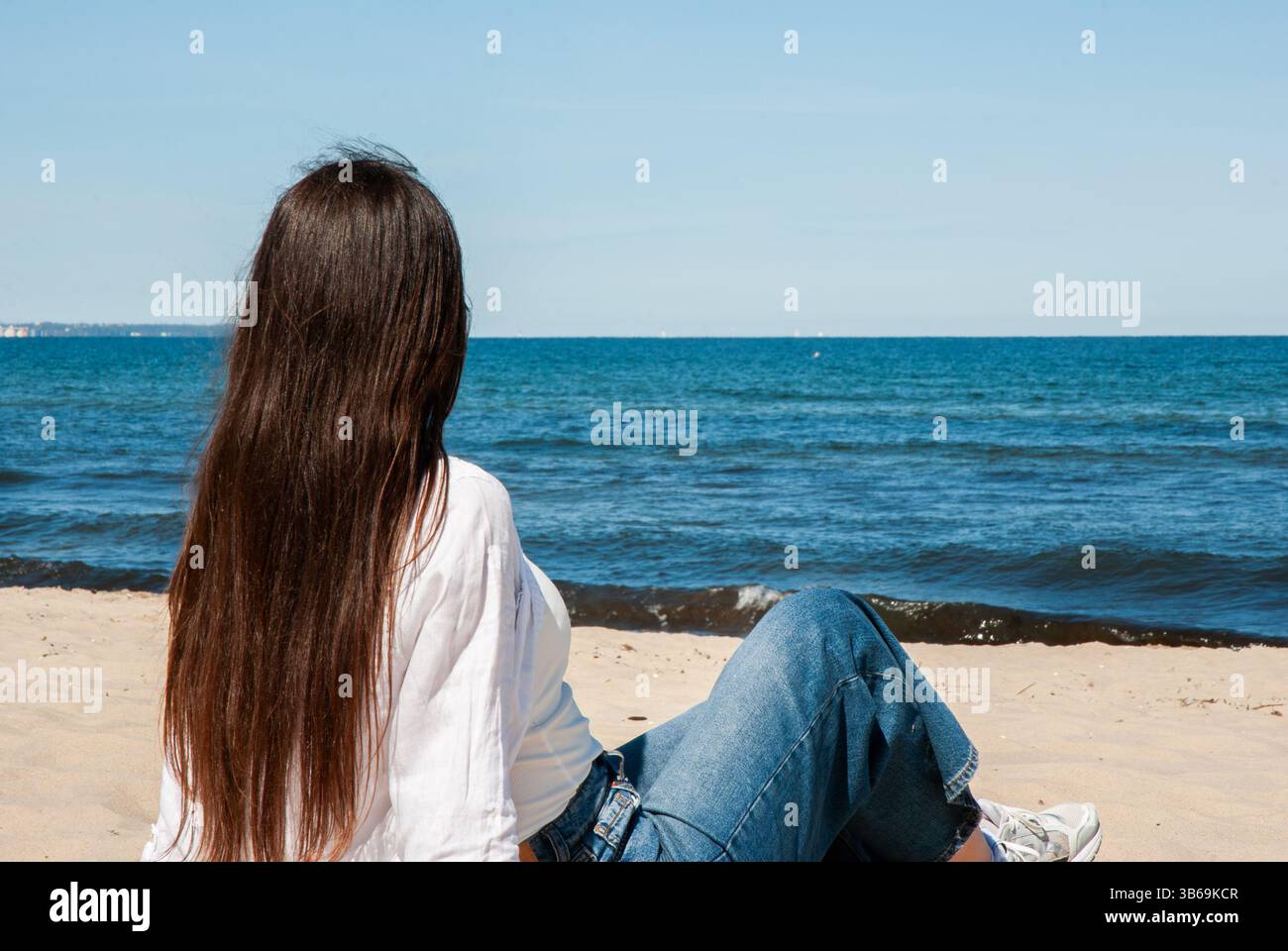Giovane donna caucasica con lunghi capelli bruna che si rilassa su una spiaggia di sabbia indossando jeans informali e una camicia bianca, guardando l'oceano blu cristallino. Digita Foto Stock