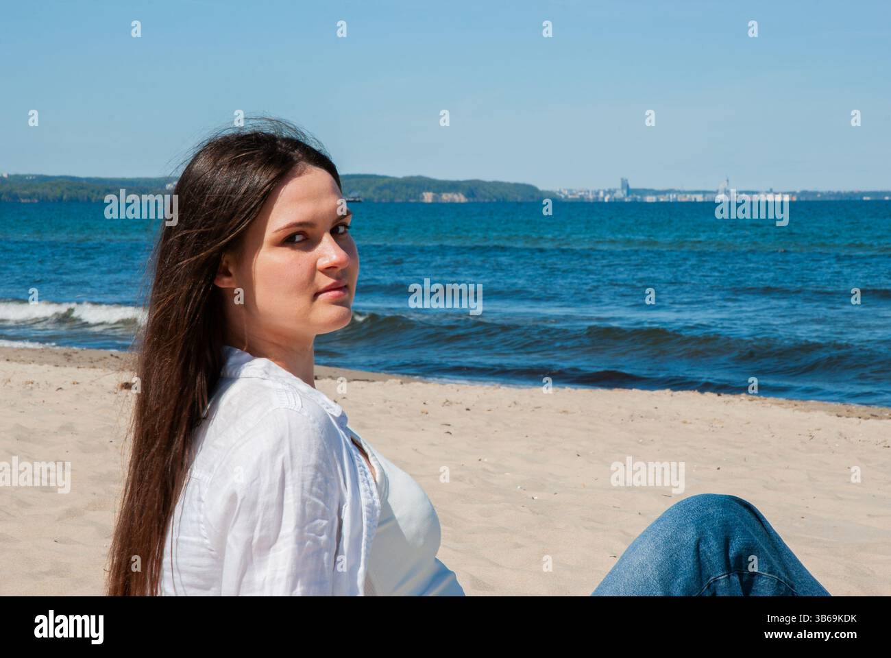 Giovane donna caucasica con lunghi capelli scuri che si rilassa su una spiaggia sabbiosa, godendo la giornata di sole con una vista distante dello skyline della città dall'altra parte dell'acqua. Foto Stock