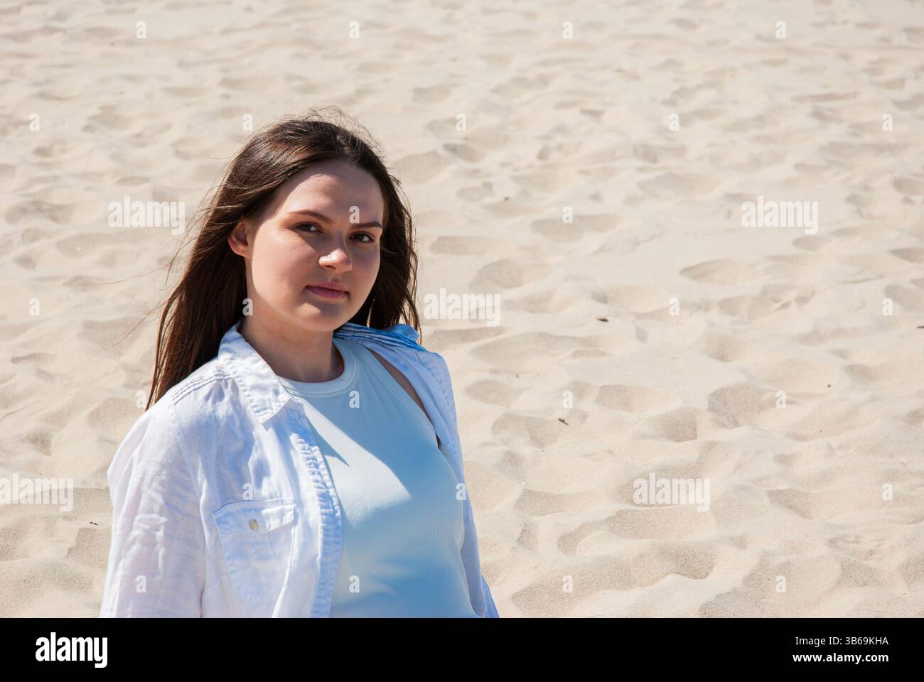 Giovane donna caucasica che si gode una giornata di sole su una spiaggia sabbiosa, indossando una giacca bianca e una parte superiore azzurra, sotto un cielo azzurro. Stile di vita dei nomadi digitali Foto Stock