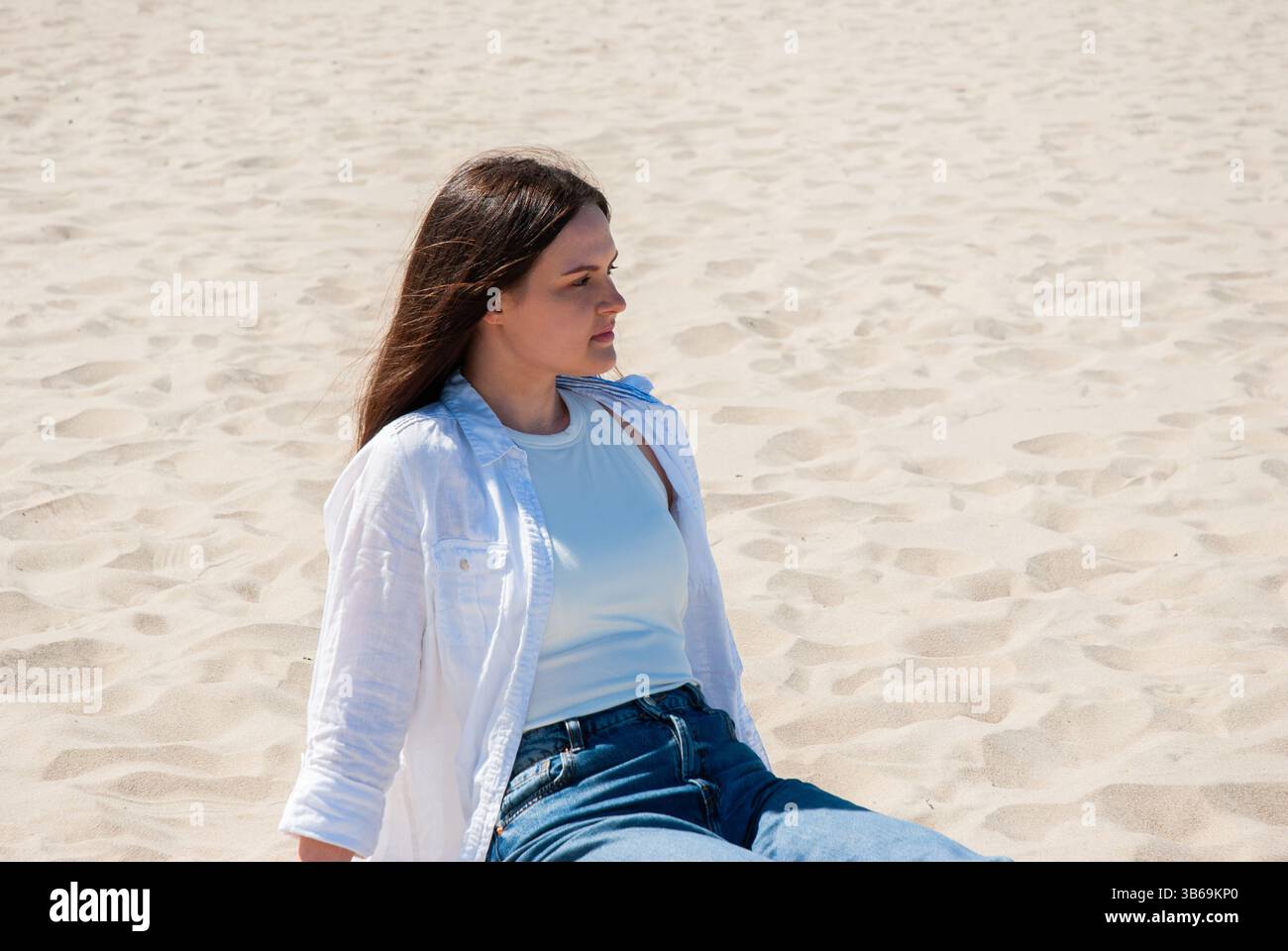 Giovane donna caucasica che si gode una giornata di sole in spiaggia con un abbigliamento casual che include una camicia bianca e jeans, seduta rilassata sulla spiaggia sabbiosa. Cifra Foto Stock