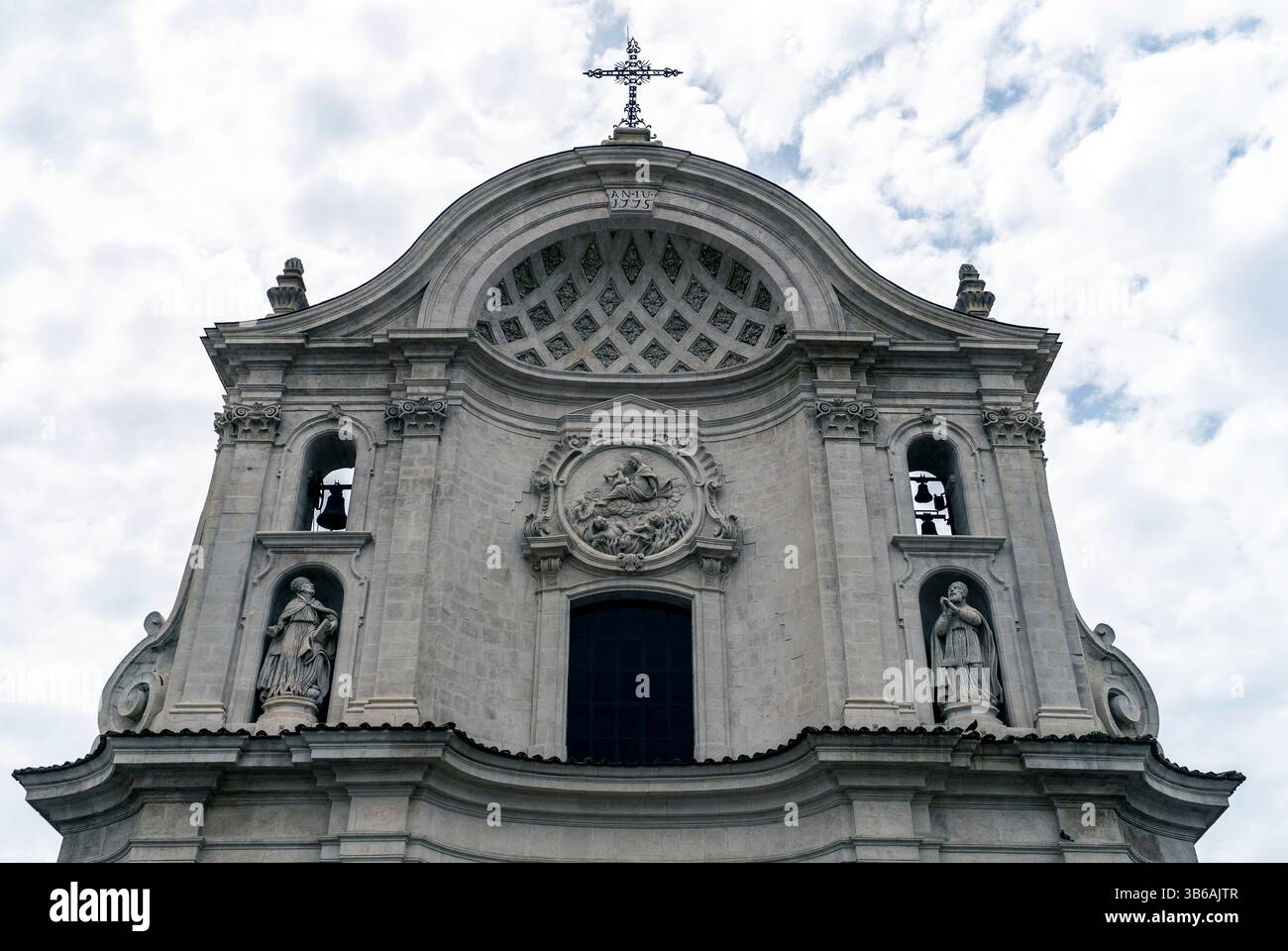 Una vista dettagliata della facciata di una chiesa con elementi architettonici ornati, statue e una croce in cima alla cupola, adagiata contro un cielo nuvoloso. Foto Stock