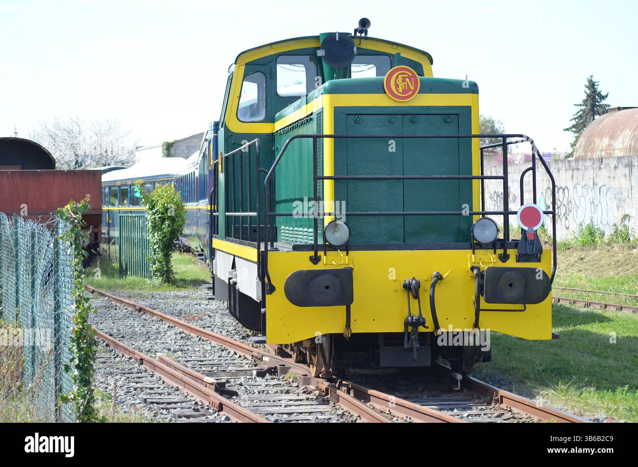 Locomotiva diesel d'epoca verde e gialla parcheggiata su binari ferroviari circondata da vegetazione lussureggiante e scenari industriali, che mostrano il fascino di Foto Stock