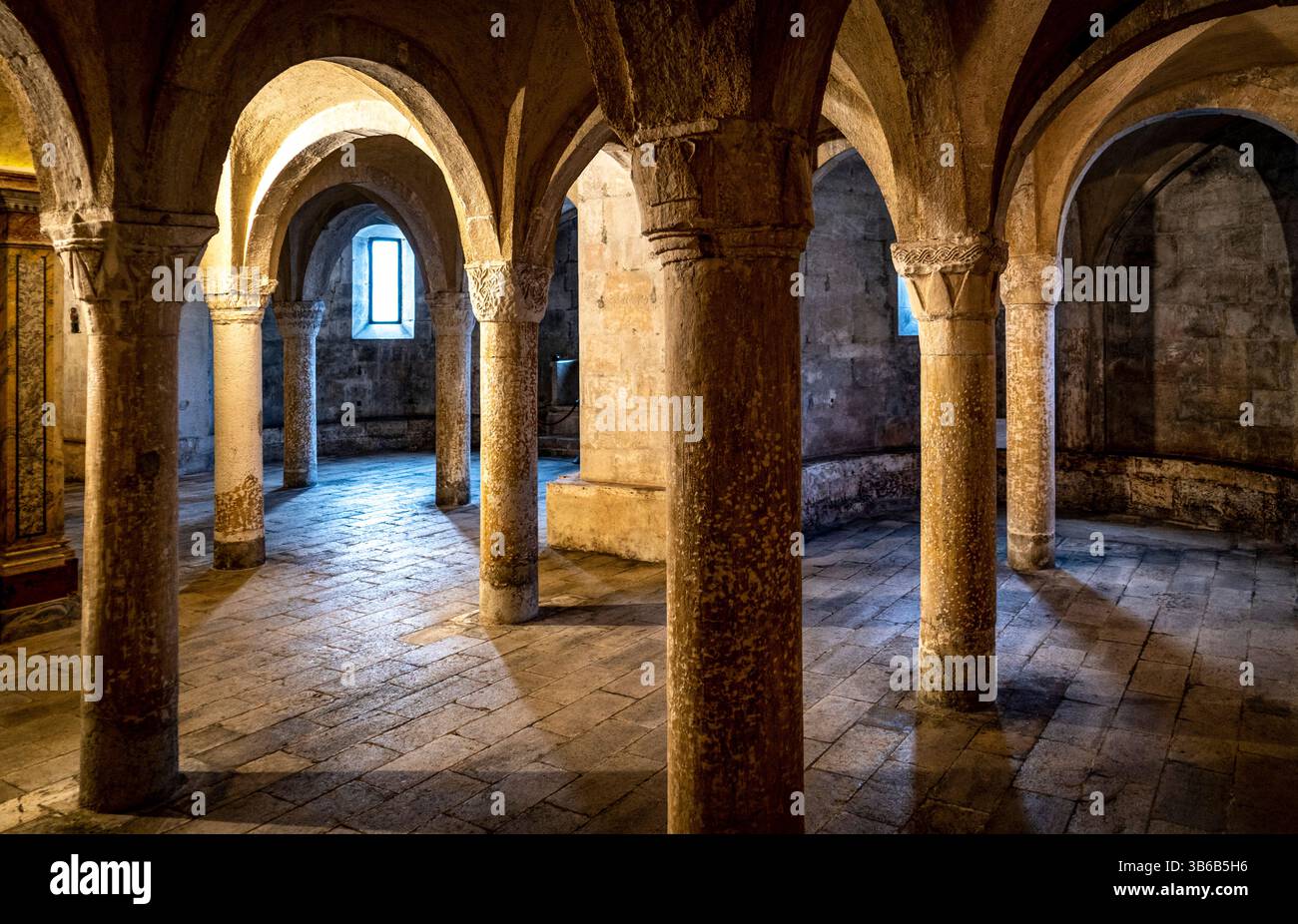 Un antico interno in pietra caratterizzato da colonne ad arco e un pavimento strutturato. La luce soffusa filtra attraverso le finestre, illuminando lo spazio e creando un ambiente sereno Foto Stock