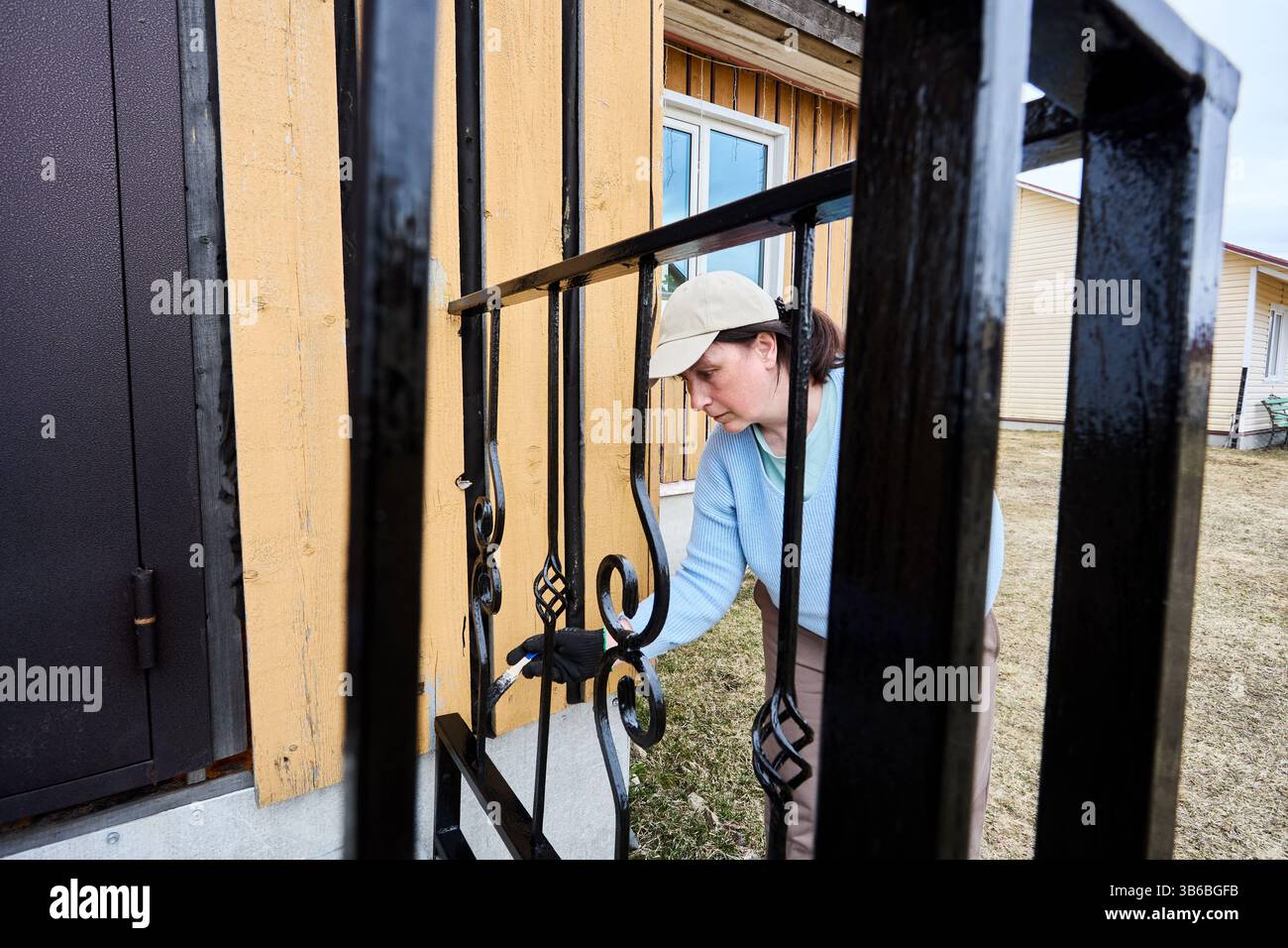 Appoggiandosi vicino a ringhiere in metallo, l'attento pittore utilizza una spazzola per coprire ogni curva e angolo, godendosi il processo di rinfrescante ingresso del portico. Foto Stock
