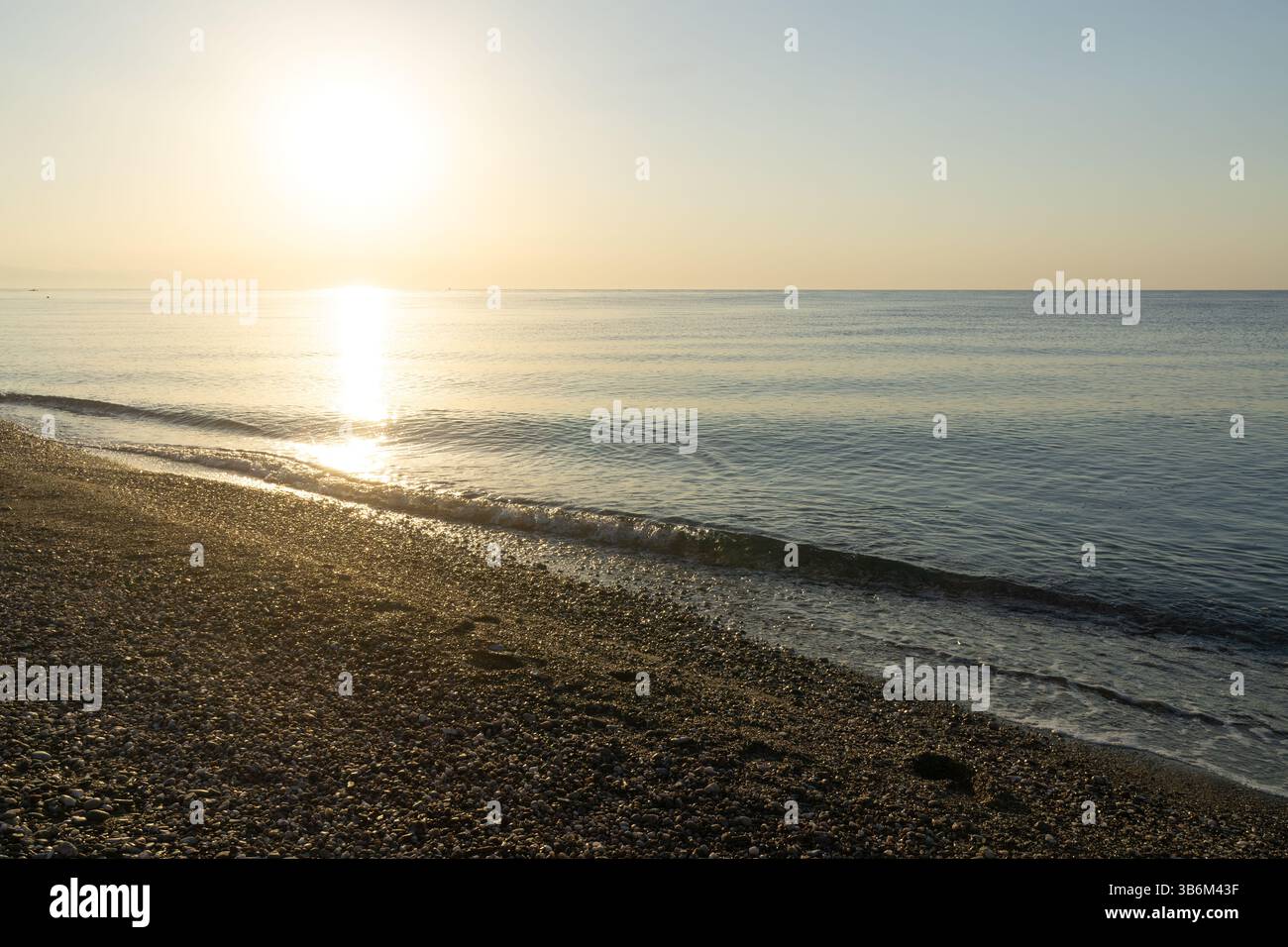 Onde dolci che lavano una spiaggia di ciottoli durante un'alba dorata su un mare calmo Foto Stock