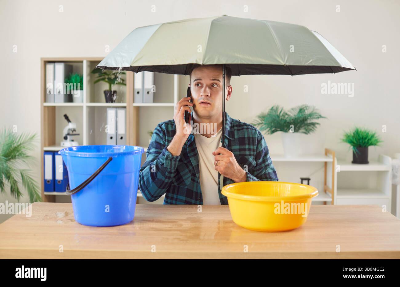 Un uomo preoccupato seduto sotto l'ombrello chiama il servizio di riparazione mentre l'acqua fuoriesce nel secchio e nel lavandino. Foto Stock